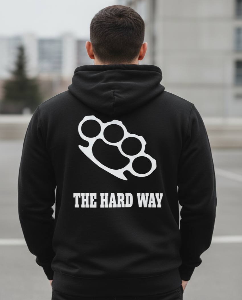 A man viewed from behind wearing a black hoodie with a white brass knuckle graphic and the text 'THE HARD WAY' printed on the back. The background is a blurred outdoor urban setting.