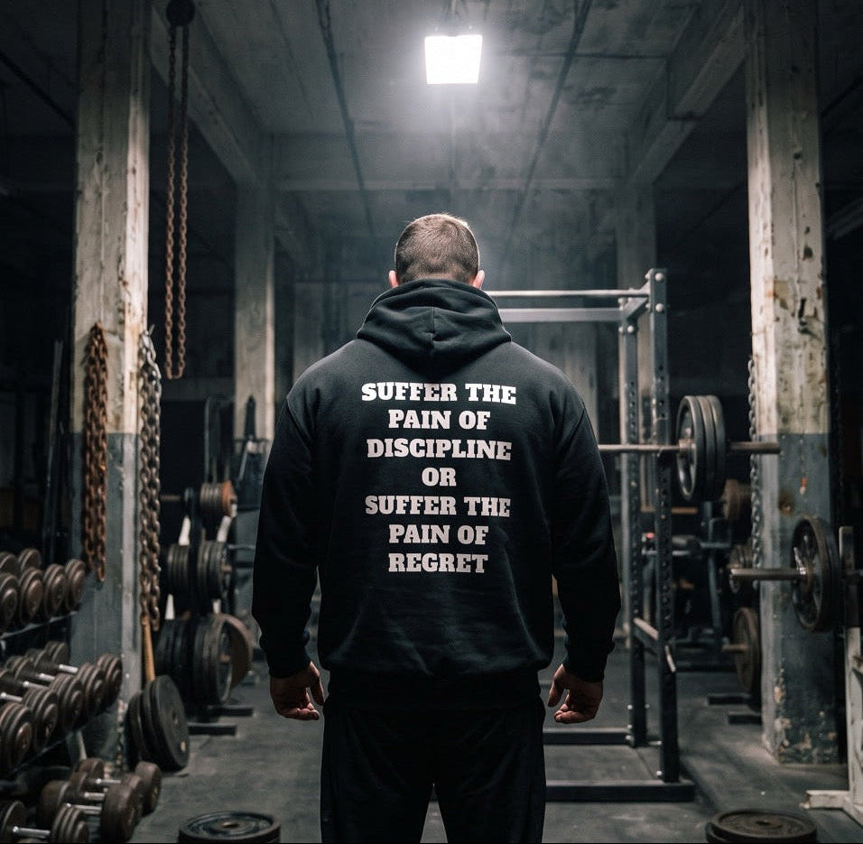 A man viewed from behind in a dimly lit gym, wearing a black hoodie with the motivational text 'SUFFER THE PAIN OF DISCIPLINE OR SUFFER THE PAIN OF REGRET' printed in white on the back.