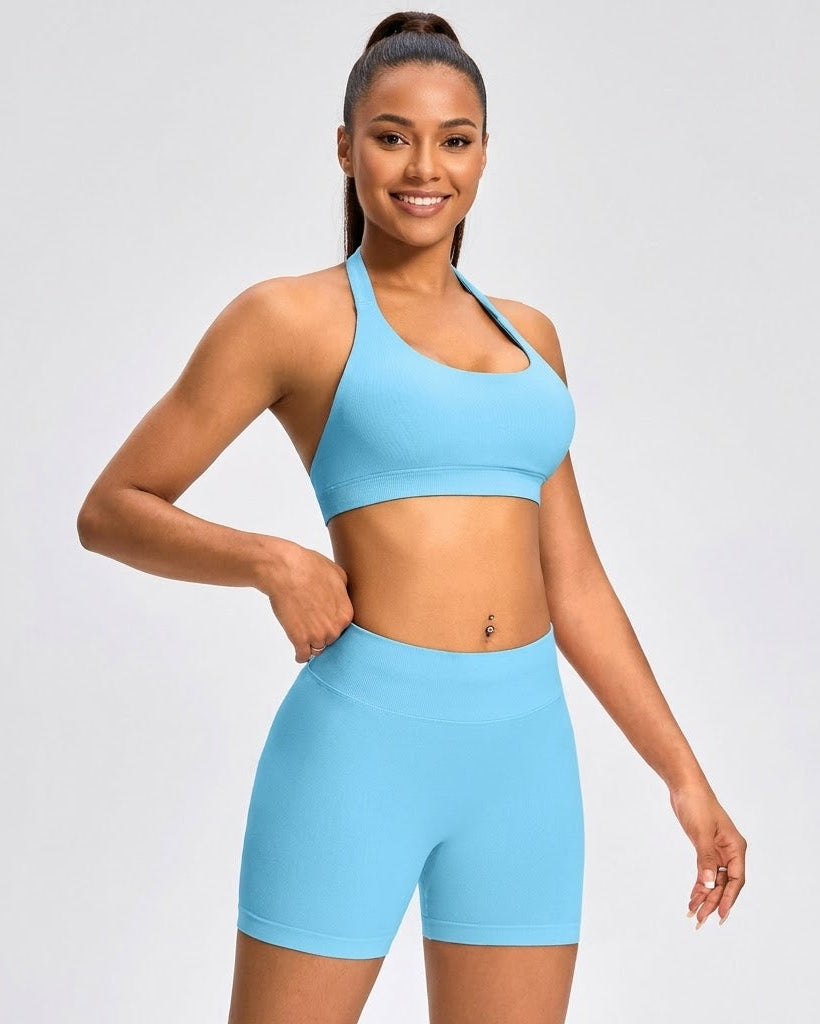 A woman modeling a light blue athletic set consisting of a sports bra and matching high-waisted shorts against a light gray studio background.