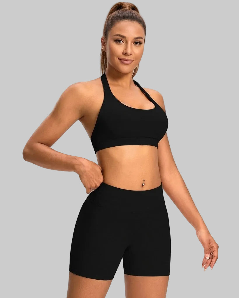 A woman modeling a matching black activewear set consisting of a halter-neck sports bra and high-waisted biker shorts, photographed against a light gray studio background.