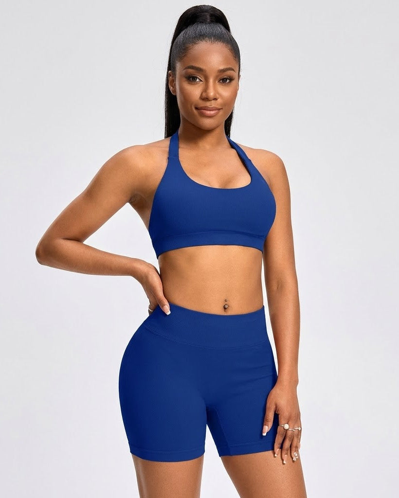 A woman modeling a matching royal blue activewear set consisting of a halter-neck sports bra and high-waisted biker shorts, set against a plain light gray studio background.