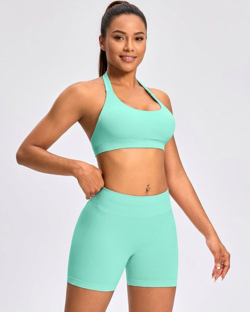 A woman modeling a matching mint green athletic set consisting of a sports bra and high-waisted biker shorts against a light gray studio background.