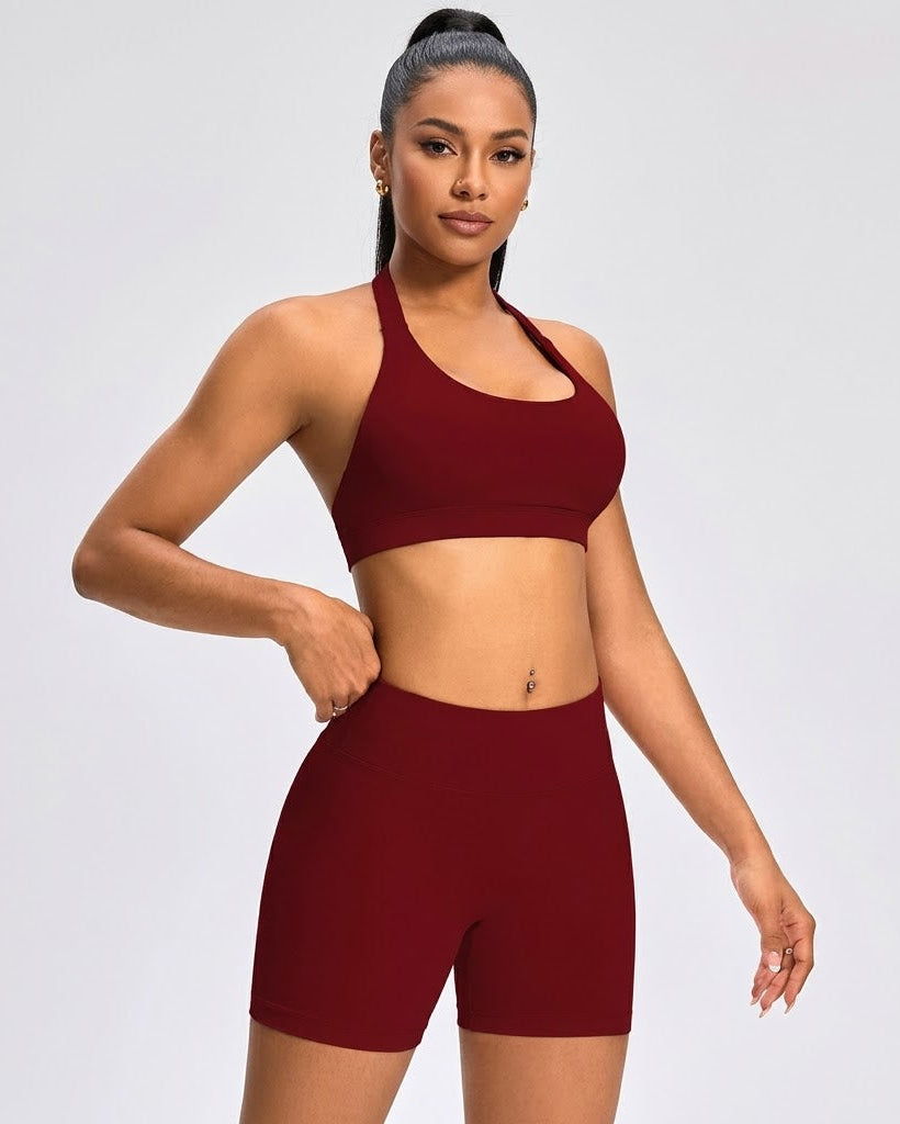 A woman modeling a matching burgundy activewear set consisting of a halter-neck sports bra and high-waisted biker shorts, set against a solid light gray background.