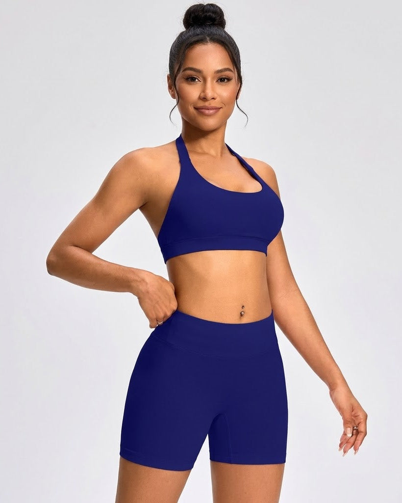 A woman modeling a matching royal blue activewear set consisting of a sports bra and high-waisted biker shorts, photographed against a light gray studio background.