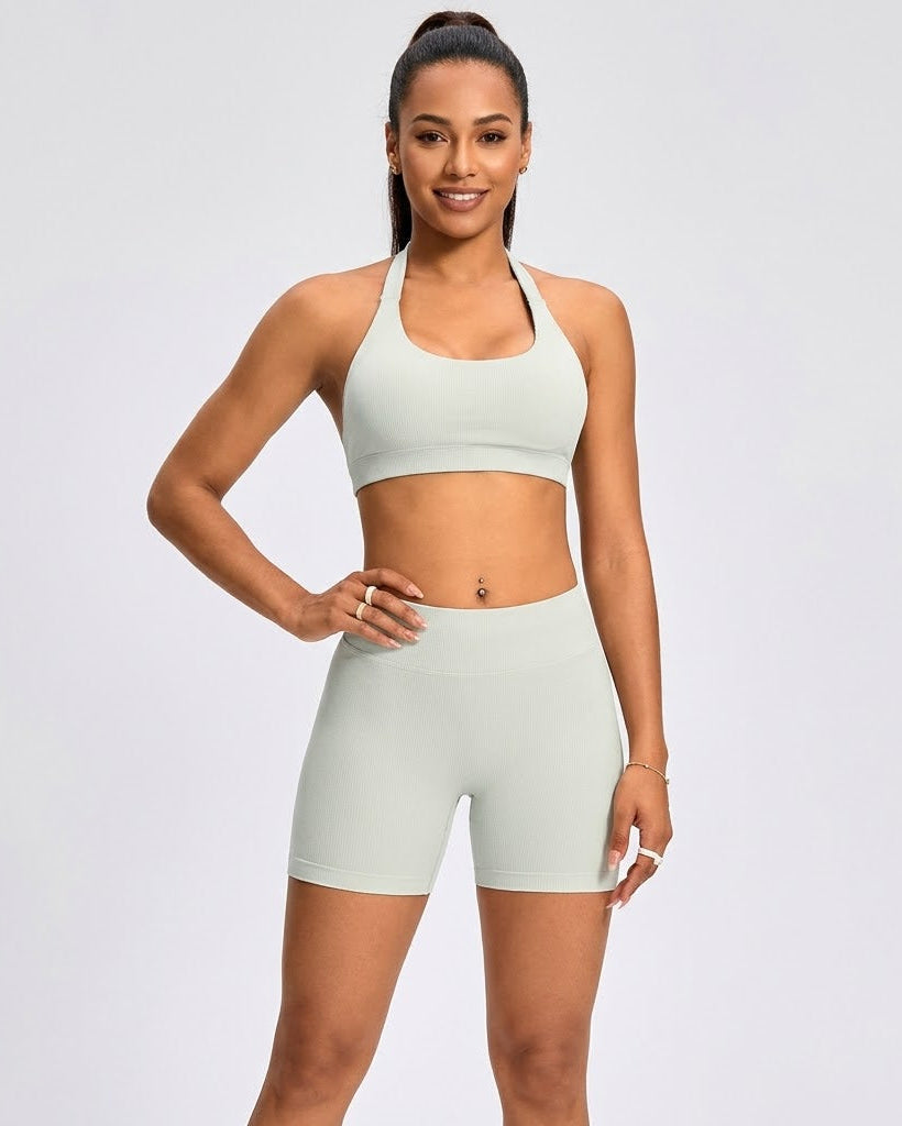 A woman modeling a matching athletic set consisting of a light sage green sports bra and high-waisted shorts against a plain light gray studio background.