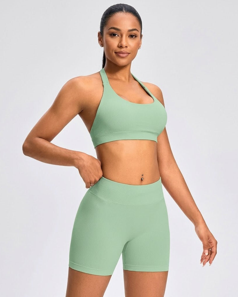 A woman modeling a matching sage green athletic set consisting of a sports bra and high-waisted biker shorts, photographed against a light gray studio background.