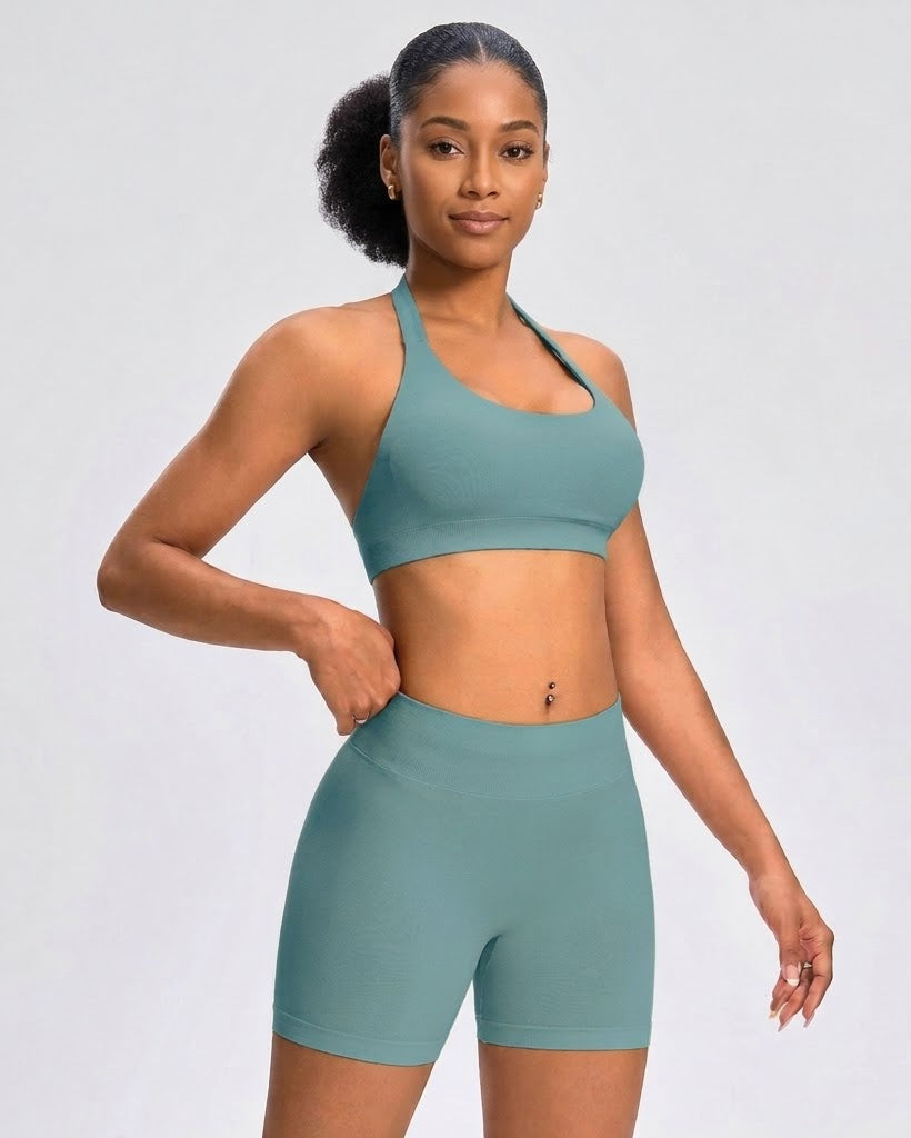 A woman modeling a matching two-piece activewear set consisting of a teal sports bra and high-waisted biker shorts, set against a plain light gray studio background.